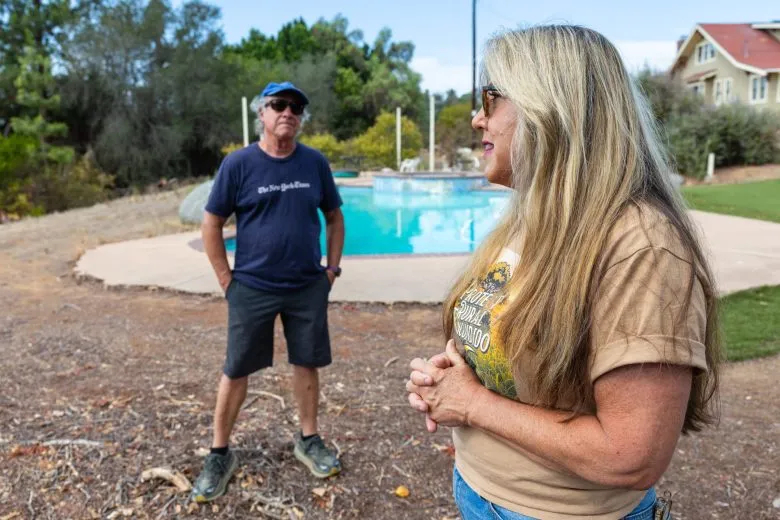 Two Escondido neighbors talking outdoors on a dry rural lot, one wearing a Protect Rural Escondido shirt, with a swimming pool, mature trees and a hillside house behind them.